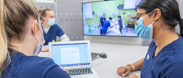 Two healthcare professionals with masks discussing work in front of a laptop, with a screen in the background displaying an ongoing medical training or simulation session.